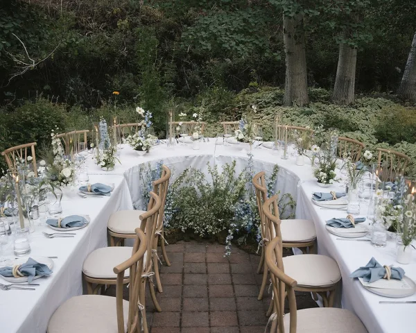 Reception tablescape with outdoor reception tables, white linens, blue napkins, bud vases and candle cylinders on a brick patio amid greenery