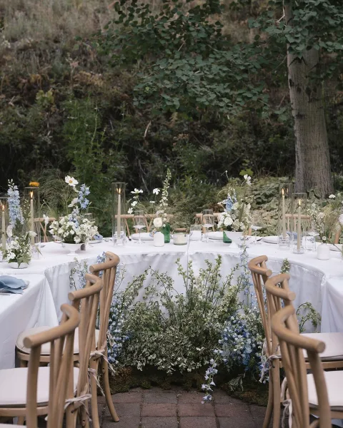 Reception tablescape with an outdoor reception table set in white linens, blue-and-white bud vases, greenery garland, and taper candles on a brick patio