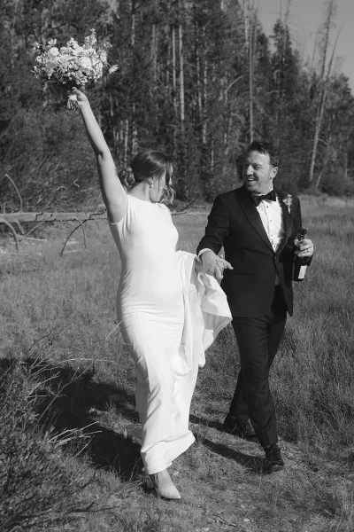 Couple portrait in a black and white wedding portrait as the bride lifts her bouquet overhead while walking with groom in a meadow near forest trees