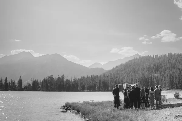 Outdoor wedding ceremony with guests gathered by a draped arch, set on a rocky lakeshore beneath pine trees and mountains