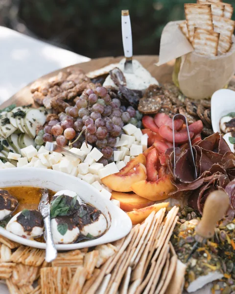 Wedding charcuterie board with cheeses, crackers, grapes, peach slices, cured meat, and a small dip bowl on a white tablecloth outdoors