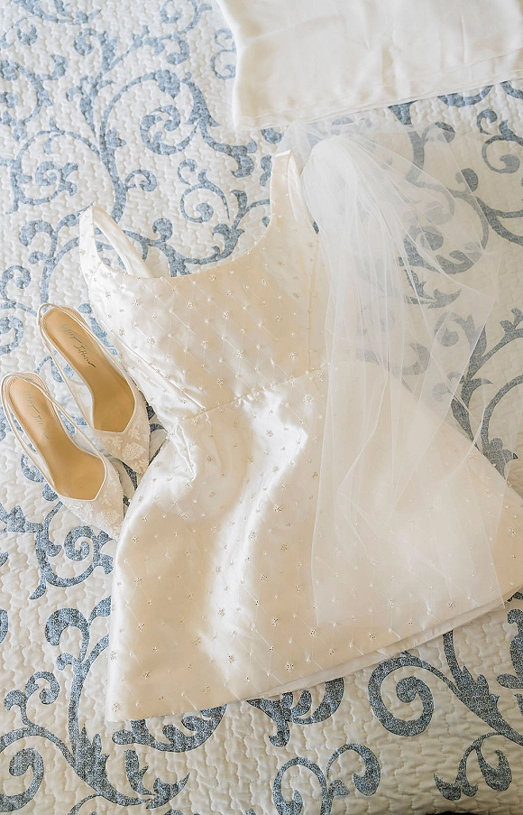 Wedding dress flatlay with tulle veil and lace bridal heels, satin bodice and beaded straps arranged on a patterned quilted bedspread