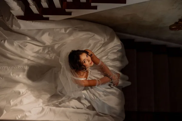 Bridal portrait of a bride on staircase, shot from above as a soft veil trails beside the banister against pale steps and wall.