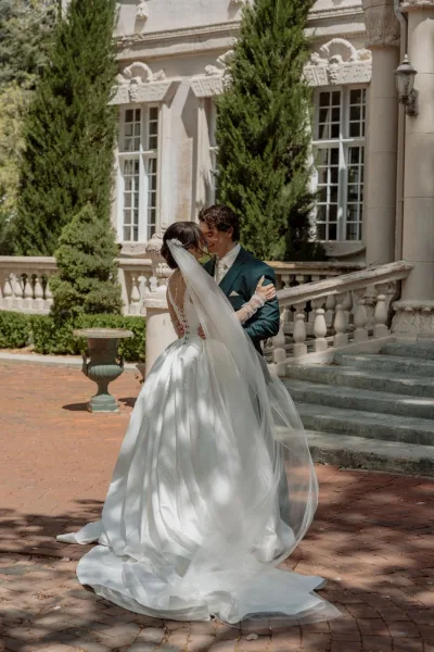 Couple portrait as a bride and groom embrace, her long veil trailing on stone steps before a mansion facade and tall evergreens.