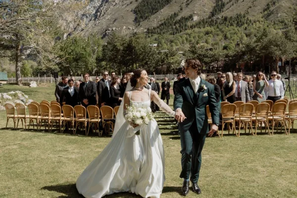 Ceremony recessional with the couple holding hands, the bride carrying a white bridal bouquet as guests cheer on a grass lawn before mountains.