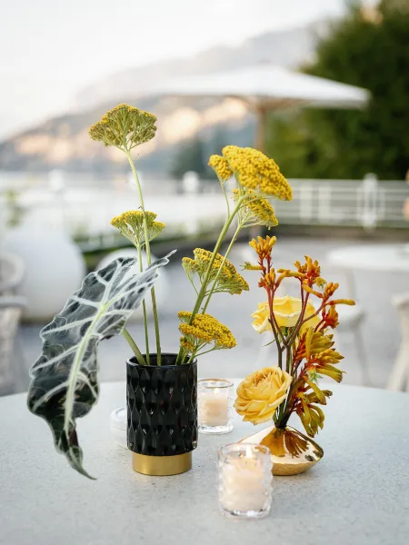Reception centerpiece with yellow wedding centerpiece buds in black and gold vases, lit by votive candles on a terrace with water and mountains