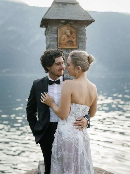 Couple portrait of bride and groom embracing, her beaded strapless gown by a lakeside with mountain reflections under open sky
