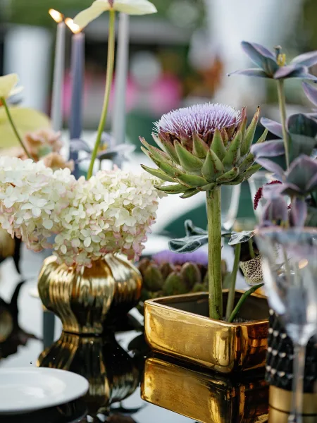 Wedding centerpiece with an artichoke wedding centerpiece in gold vases, taper candles, and champagne flutes on an outdoor table with greenery behind