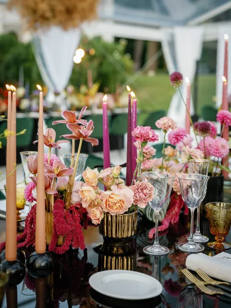 Reception tablescape on a black wedding table with pink and burgundy florals, gold vases, taper candles, and amber votives under a draped tent
