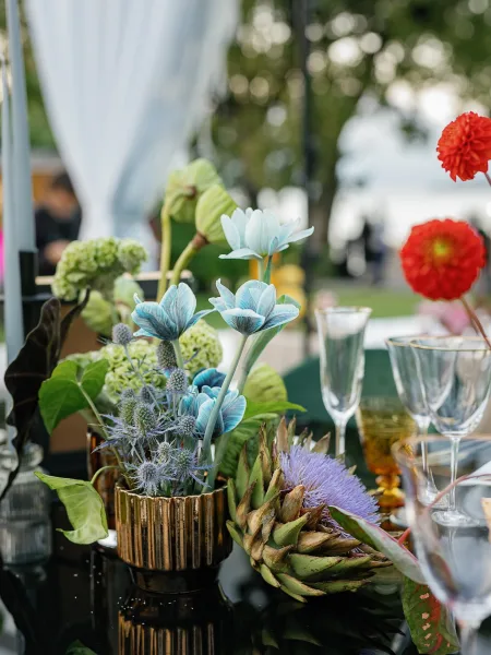 Reception centerpiece with wedding floral centerpiece in a gold vase, blue and red blooms with greenery, set among goblets and blurred guests outdoors