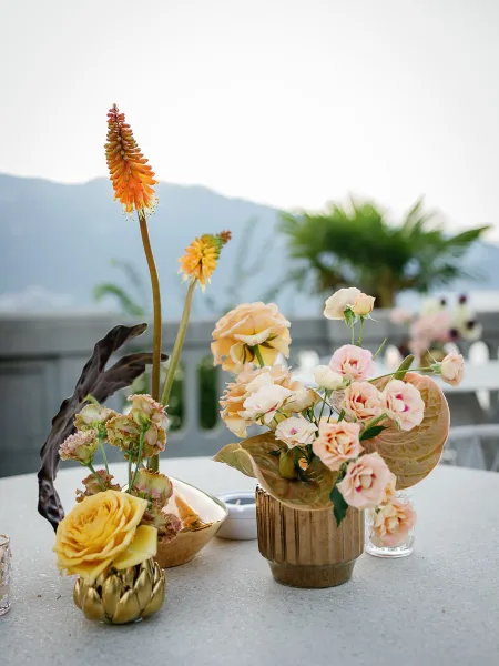 Wedding centerpiece with roses and anthurium leaves in gold vases and bud vases on linen, set on an outdoor terrace with mountains beyond