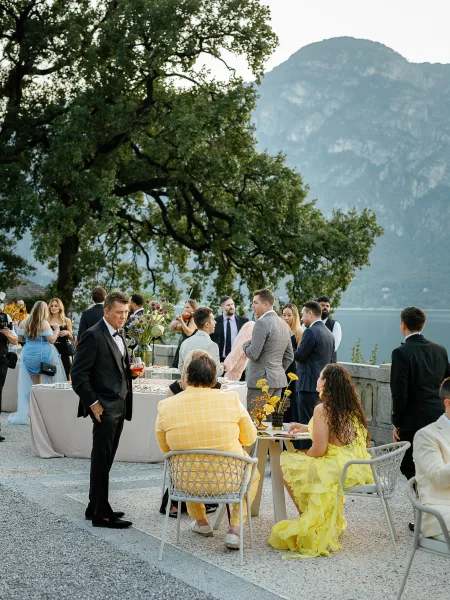 Wedding cocktail hour with wedding guests mingling around linen high-top tables, cocktails and florals on a stone terrace by a lake and mountains