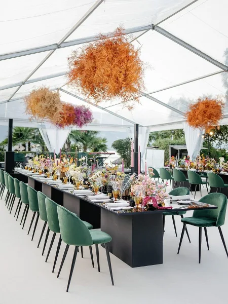 Reception tablescape with long banquet table wedding setup, green velvet chairs, colorful taper candles, and hanging florals under a clear tent in a palm garden