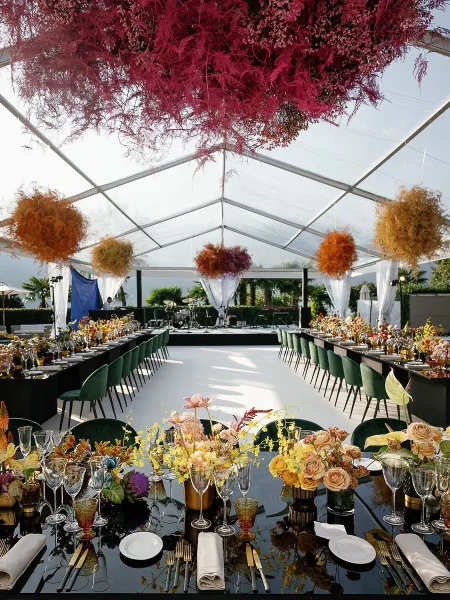 Reception tablescape with a long banquet table setup under a clear top tent, colorful florals, amber goblets, and green velvet chairs