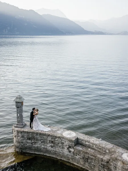 Wedding kiss on a stone pier, bride in flowing gown and veil embracing groom in tuxedo above rippling lake with mountain backdrop