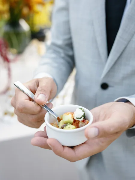 Wedding cocktail hour food in appetizer cups held by a guest in a gray suit jacket with black tie, with blurred florals in outdoor light
