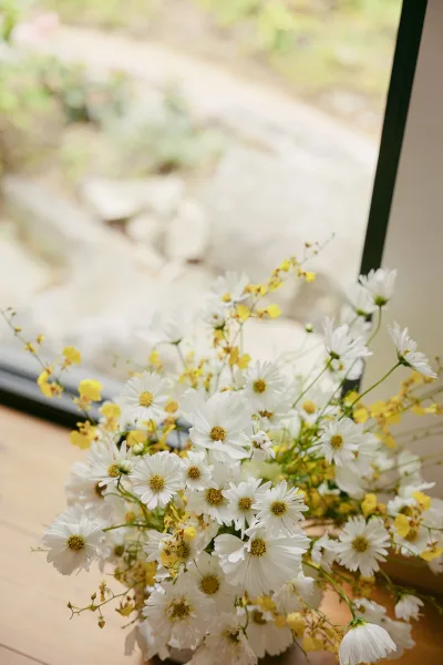 Bouquet close-up of a white daisy bouquet with yellow flowers, set on a wood floor beside a window frame and garden path.