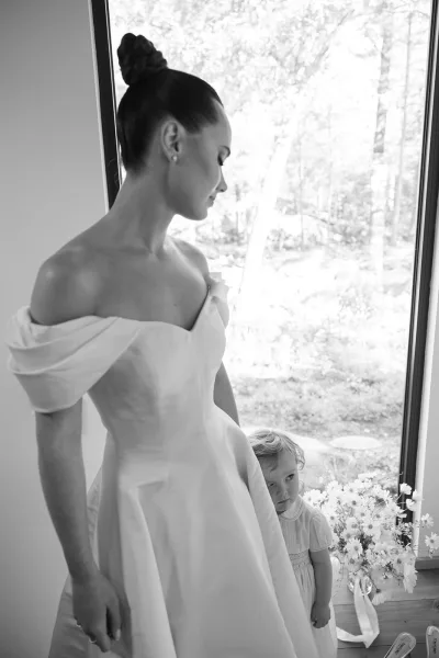 Bride with flower girl share a candid wedding moment by a bright window with garden trees, the bride cradling a simple bouquet.