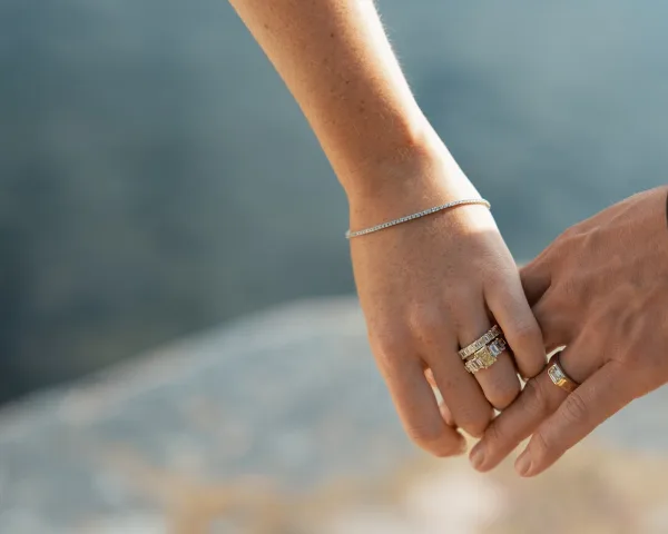 Wedding rings in a bridal ring stack sparkle beside a delicate bracelet on stone ground, with soft water reflections in the background.