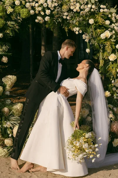 Couple portrait in a wedding dip pose beneath a floral arch on sandy ground, capturing a romantic moment.
