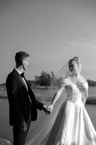 Black and white couple portrait of a bride and groom holding hands, her bridal veil framing them beside a rocky shoreline of water and trees.