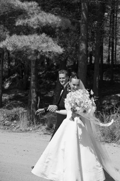 Couple portrait of a bride and groom walking hand in hand, the bride holding a bouquet along a forest dirt path bordered by pine trees and tall grass.
