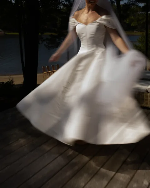 Bridal portrait of a bride in a twirling bride pose on a lakeside wooden deck, her veil lifting softly amid trees and sand.