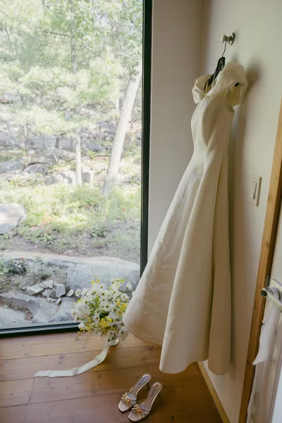 Wedding dress, an ivory wedding gown paired with a bridal bouquet, by a large window overlooking forest trees and rocks.