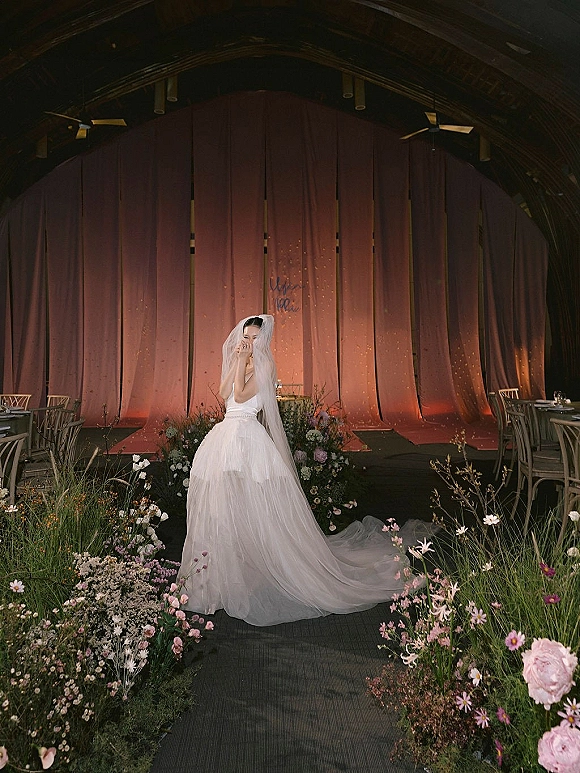 Bridal portrait of a bride with long veil over her face, standing by a flower-lined aisle runner in a warm-lit indoor hall with stage backdrop