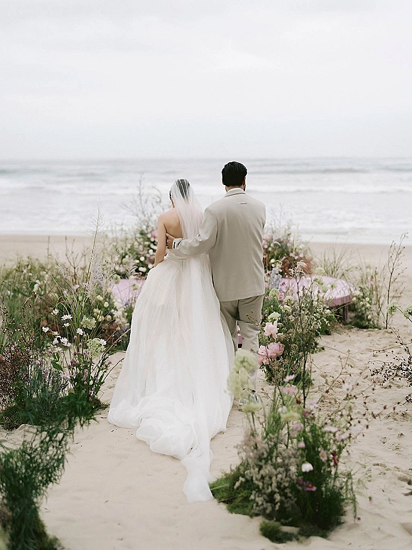 Couple portrait at a beach wedding, bride and groom from behind embracing, long veil and strapless dress facing the ocean horizon
