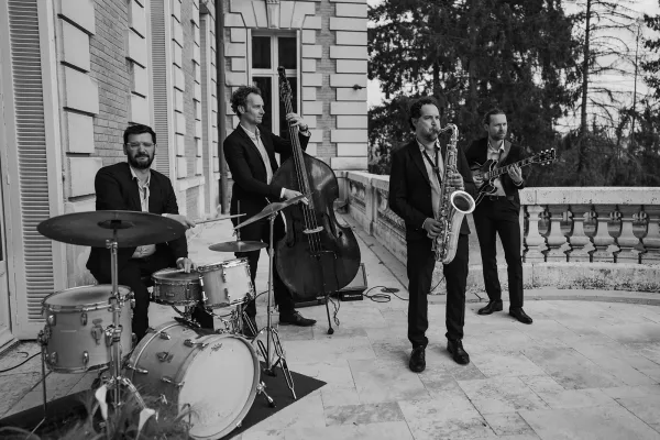 Live wedding band performs as a jazz wedding band, featuring a saxophone, on a stone terrace outside a grand mansion with trees beyond.