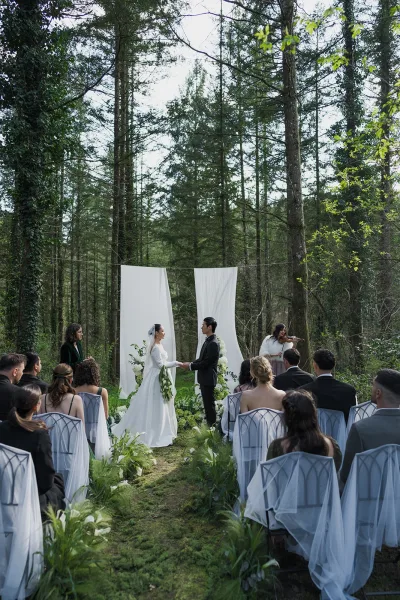 Ceremony moment at an outdoor wedding ceremony as the bride and groom exchange vows beneath forest trees on mossy ground, a violin nearby.