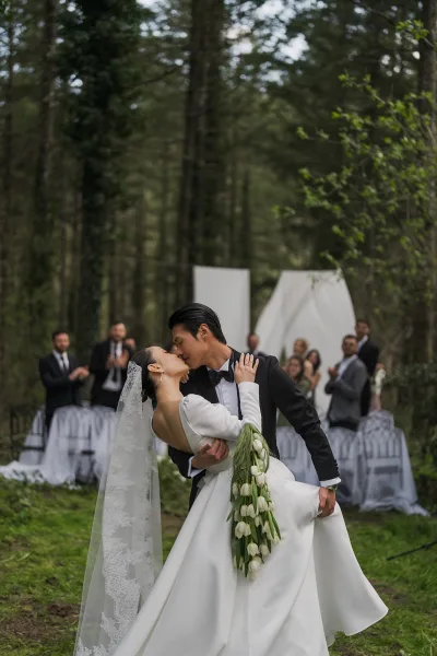 Wedding kiss portrait capturing a ceremony kiss moment, bride in a lace veil and groom embraced amid forest trees and green lawn.