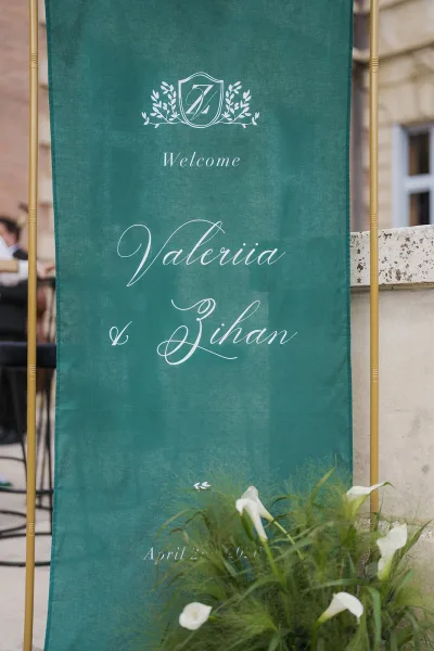 Wedding welcome sign in emerald green on a gold stand, set by a stone wall and greenery on an outdoor terrace.