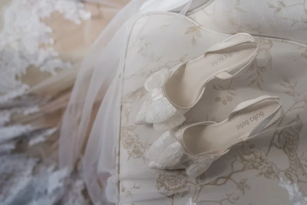 Bridal shoes in ivory lace with closed toes and scalloped edges, resting on a floral upholstered chair.