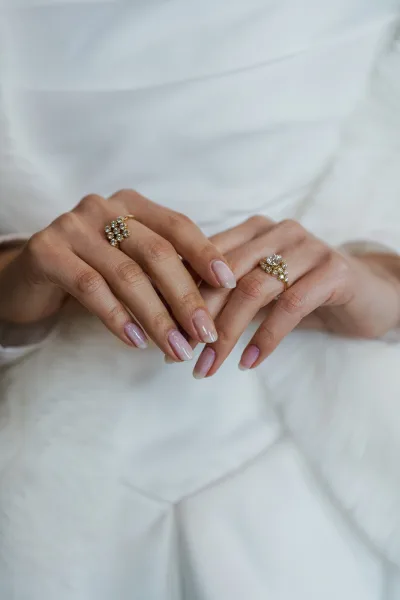 Bridal manicure with sheer pink wedding nails on almond tips, hands resting on bridal gown fabric, accented by delicate rings.