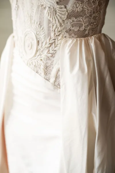 Wedding dress close-up showing a lace wedding dress bodice with embroidered floral applique and pleated satin skirt detail on a neutral wall