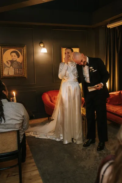 Ceremony moment as the couple share a vow reading, the bride holds a vow book before a dark paneled wall and velvet sofa in an intimate setting.