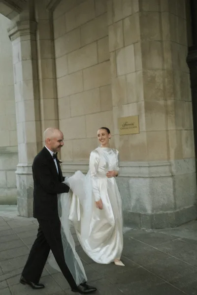 Bride and groom walking in a classic walking wedding photo pose, her veil trailing as they pass a stone archway with columns along the sidewalk.