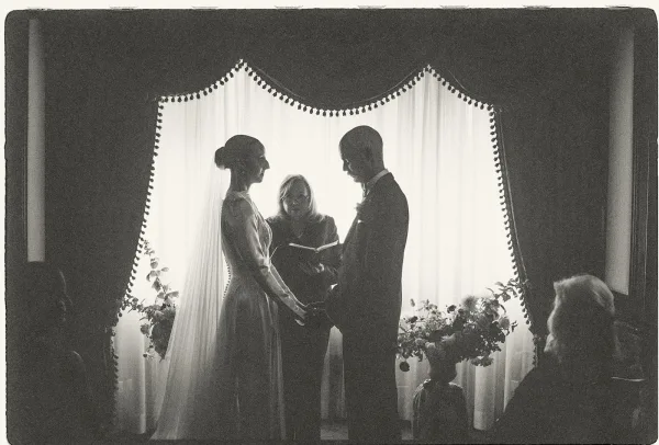 Ceremony moment with a couple exchanging vows by window light, draped curtains behind, the bride’s veil softly lit in this classic indoor scene.