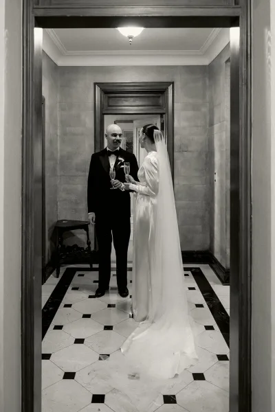 Couple portrait in a champagne toast moment, the bride's long veil trailing as they smile close together before taking a celebratory sip.