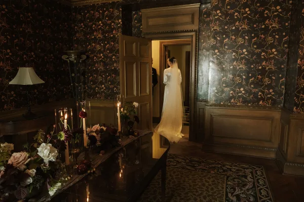 Candlelit bridal portrait of a bride in a doorway, the scene warmed by candles and framed by floral wallpaper and wood paneling.