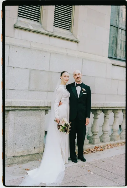 Couple portrait of bride and groom standing, her satin long-sleeve gown flowing as they pose by a stone balustrade sprinkled with fallen leaves.
