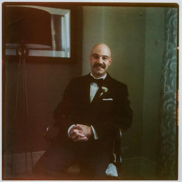 Groom portrait of a black tie groom with a boutonniere, posed before a neutral wall with framed artwork and a soft curtain.