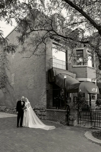 Couple portrait of a bride and groom outside, her long veil trailing as they stand by a brick building and iron fence along a tree-lined sidewalk.