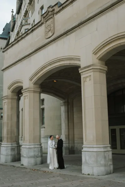 Couple portrait, a wedding portrait under stone archways, the bride in a long-sleeve gown beside the groom, framed by a historic facade.