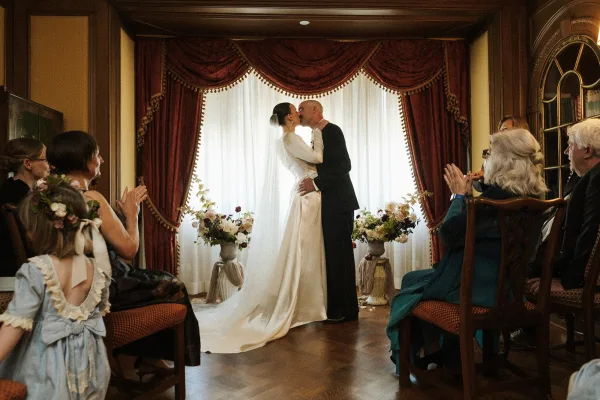 Ceremony kiss moment as the bride and groom embrace at an indoor wedding ceremony, framed by rich red drapery.