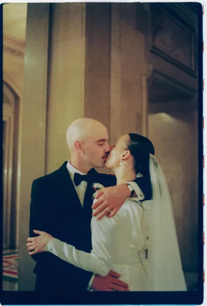 Wedding kiss portrait of a bride in a long sleeve gown and groom, her long veil trailing beneath stone columns and an ornate archway indoors.