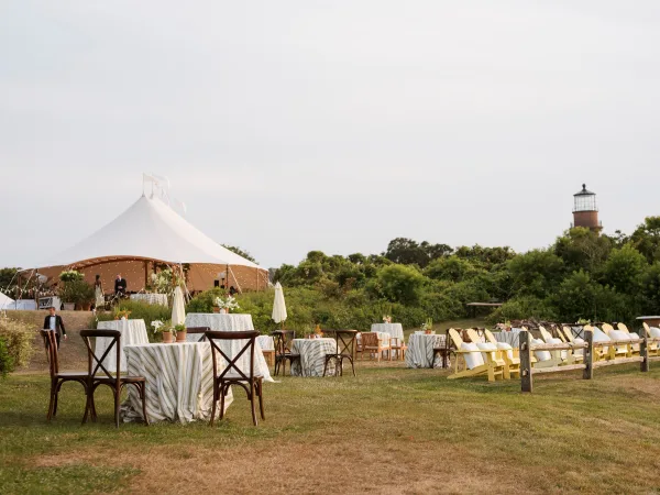 Outdoor reception setup under a sailcloth wedding tent with string lights, round tables, patio umbrellas, and a lighthouse beyond the lawn