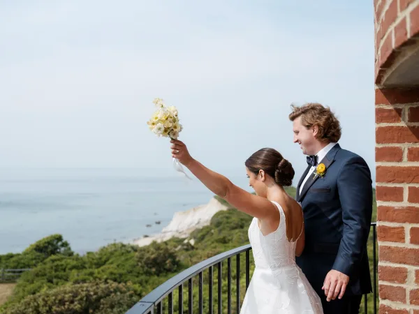 Couple portrait on a wedding balcony, bride holding bouquet up beside groom in tuxedo, with ocean cliffs and sky behind